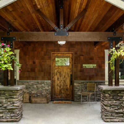 Custom covered front entry with exposed timber beams, decorative steel brackets, natural wood ceiling, stone columns, and rustic wood front door.
